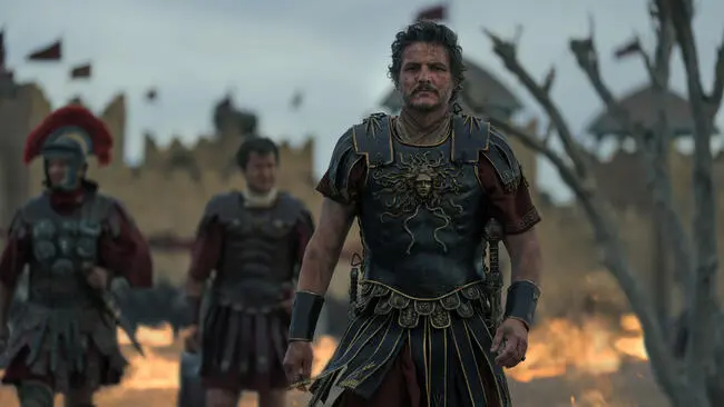 Pedro Pascal como comandante romano en un campo de batalla, vestido con armadura detallada, con soldados en el fondo y llamas crepitantes tras la destrucción